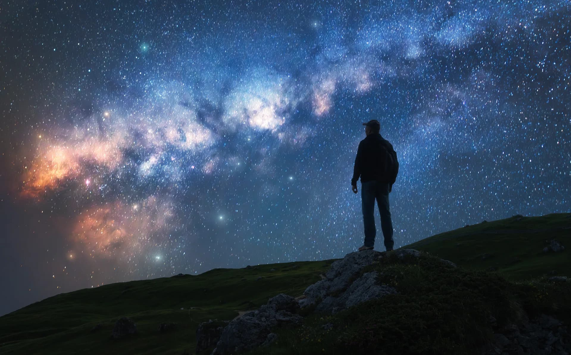 A single person standing on a rocky outcrop looking up at a vast, starry night sky — quiet contemplation and philosophical wonder