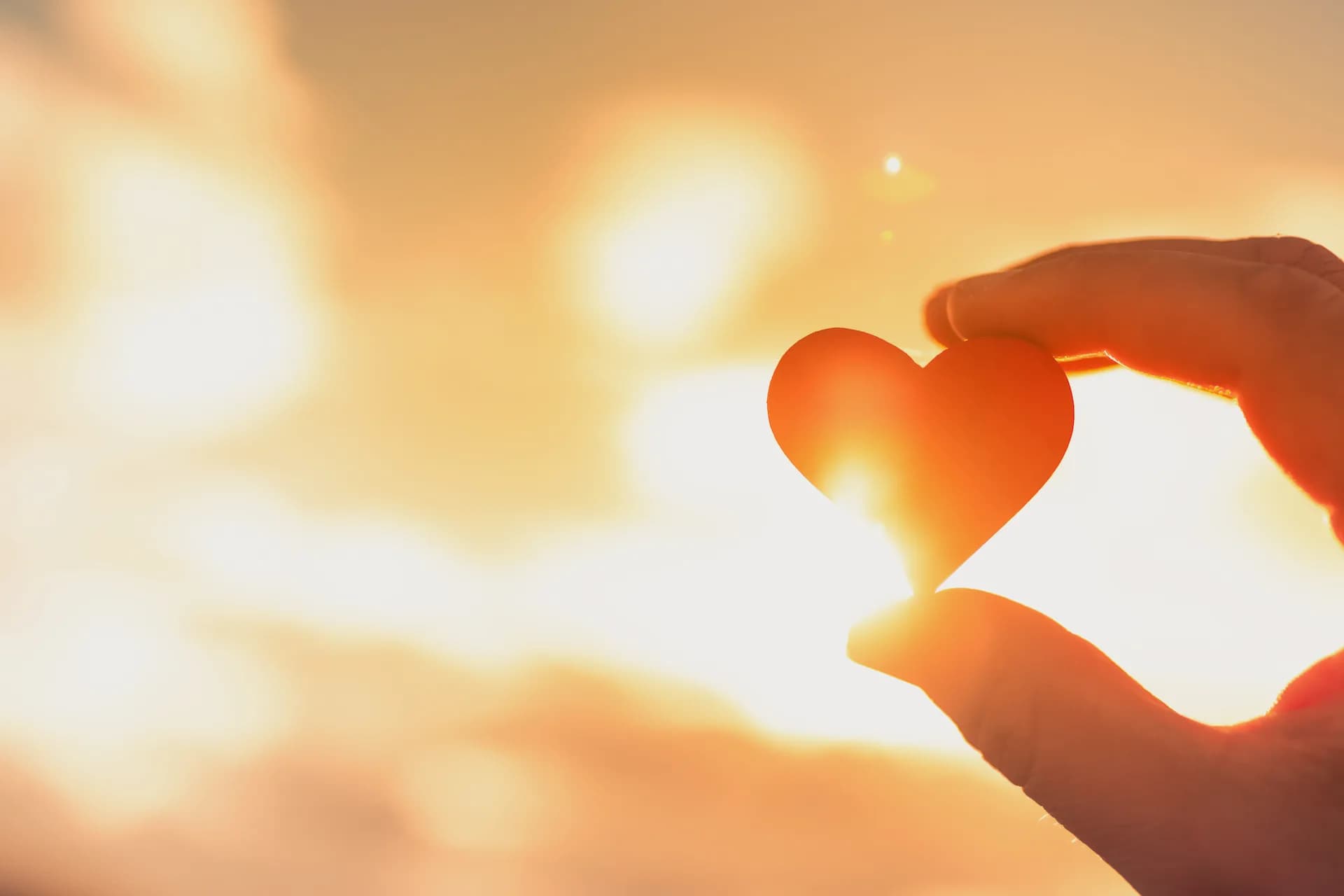A hand holding a paper heart up against a golden sunrise sky