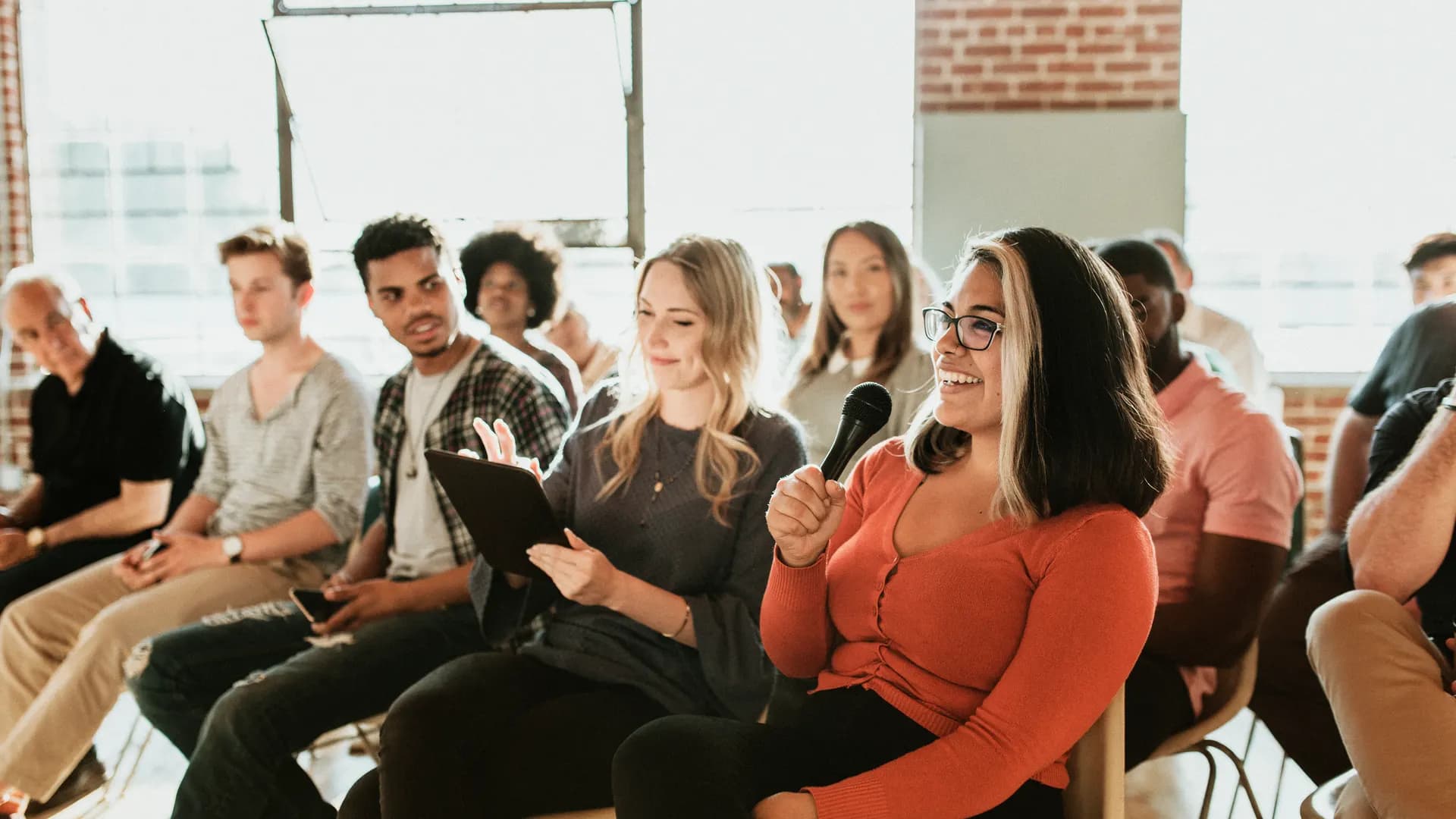 A room of people listening at a live training session, one of them speaking into a microphone