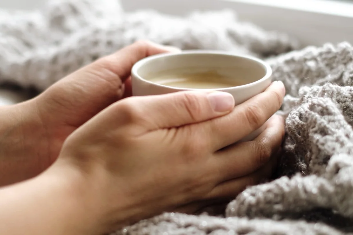 Las manos de una persona descansando alrededor de una taza de té caliente: conectando a tierra y cuidando nuestras necesidades humanas mientras sostenemos el dolor espiritual