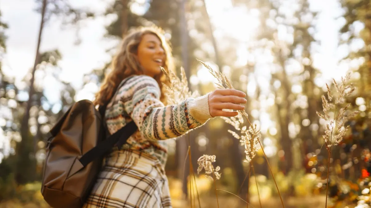 A woman walking through a sunlit forest with arms outstretched, open to noticing the beauty and energy available in the natural world around her
